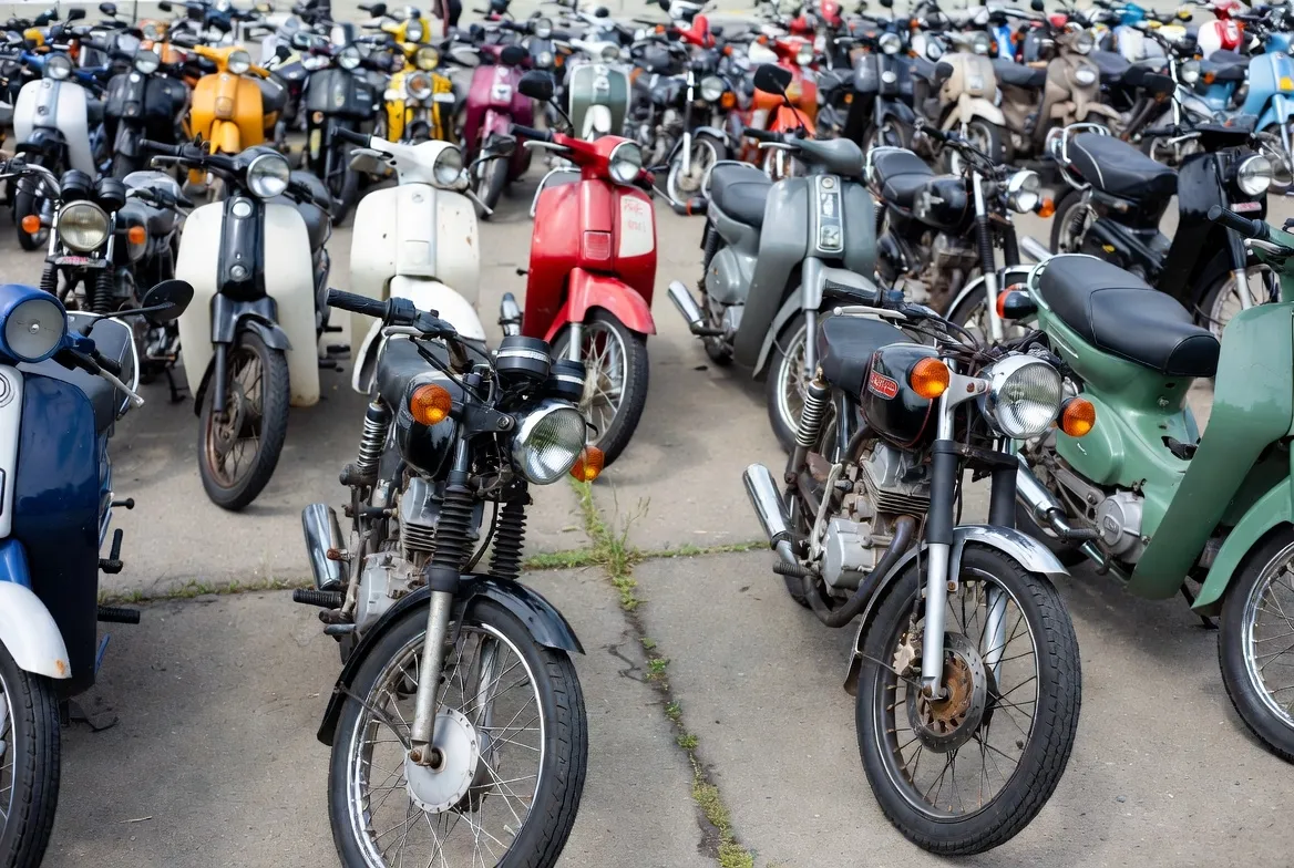 A photograph of old motorcycles in a lot, arranged randomly under overcast daylight.